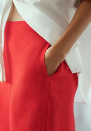 Gros plan d'une personne portant un pantalon rouge avec une main dans la poche et une chemise blanche partiellement rentrée.