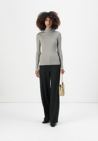 Lys grå ribbet turtleneck genser kombinert med svarte stripete vide bukser og en liten beige håndveske, stående foran en ensfarget bakgrunn.