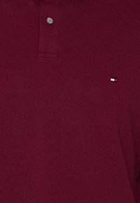 Burgundowa koszulka polo z fakturowanej tkaniny, z dwoma guzikami na klatce i małym logo w kolorach czerwonym, białym i niebieskim blisko dołu.