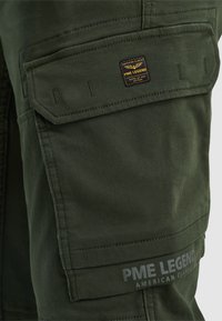 Pantalon cargo vert foncé avec une poche à rabat affichant un petit patch logo PME Legend et le texte imprimé PME Legend près de l'ourlet.