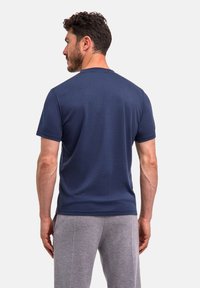 Man met kort krullend haar in een marineblauw overhemd met korte mouwen en grijze broek, staand met zijn rug naar de camera tegen een effen achtergrond.