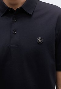 Tengerészkék póló puha pamutból, három fekete gombbal, klasszikus gallérral és egy finom logófolttal a bal mellrészen.