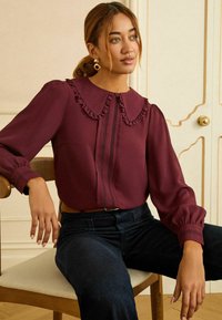 Bordeauxröd blus med rynkad krage, centrala veck och mörka detaljer på ärmarna. Buren med mörka jeans, sittande på en trästol.