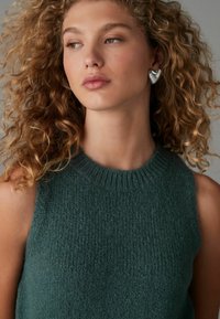 Türkisfarbener, ärmelloser Strickpullover mit rundem, geripptem Ausschnitt. Strukturiertes Material und dezenter Glanz. Das Model trägt herzförmige silberne Ohrringe.