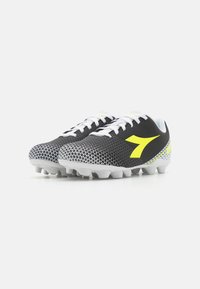 Scarpe da calcio bianche e nere con parte superiore testurizzata, logo giallo e motivo geometrico bianco sulla suola. Progettate per superfici in erba.