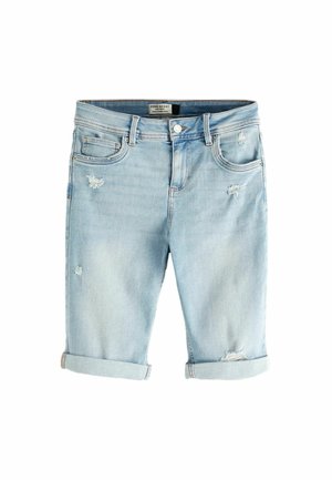 Femme portant un chemisier en dentelle blanche et un short en jean bleu clair déchiré, debout près d’une côte rocheuse avec l’océan en arrière-plan.