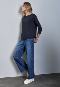 Tummansininen kolme neljännestä hihasta oleva paita yhdistettynä leveälahkeisiin sinisiin farkkuihin, joissa on koristeellisia niittejä, ja täydentänyt asua valkoisilla tennareilla tekstuurilla varustetulla pinnalla.