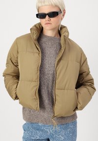 Khaki puffer takki, jossa on korkea kaulus, tikattu muotoilu, etuvetoketju ja joustavat hihansuut, kerroksittain harmaan neulotun villapaidan päällä.