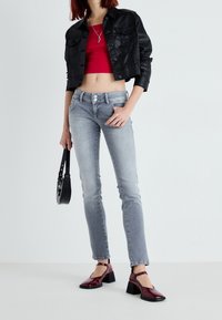 Une personne portant une veste en cuir noire courte, un crop top rouge, un jean slim gris, des chaussures à talons carrés bordeaux et tenant un petit sac à main noir.