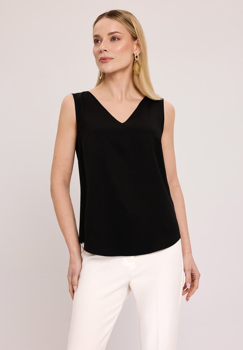 Czarny bezrękawnik z dekoltem w serek, gładką fakturą i lekko zaokrąglonym dołem, w połączeniu z białymi, dopasowanymi spodniami.