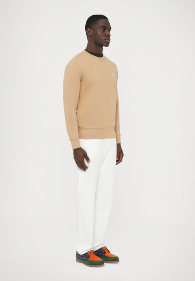 Bež pulover z okroglim izrezom, rebrastimi manšetami in spodnjim robom, kombiniran z belimi hlačami in večbarvnimi čevlji z zelenimi, oranžnimi in modrimi poudarki.