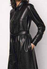 Černé kožené šaty košilového střihu s límečkem, dlouhými rukávy, předními knoflíky, kapsou na hrudi a látkovým páskem v pase, hladká textura.