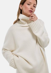 Pull à col roulé en tricot doux. Présente des poignets et un col côtelés, une coupe ample et une poche latérale. Texture lisse, design chaud et confortable.