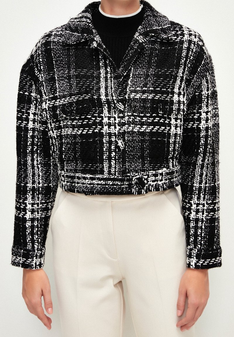 Veste courte à carreaux noirs et blancs portée par-dessus un top noir, associée à un pantalon taille haute blanc cassé, montrée du cou aux hanches.
