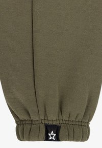 Ύφασμα ελιάς πράσινο, ελαστική μανσέτα με ρυτιδωτή υφή και μαύρη ετικέτα με λευκό λογότυπο αστεριού. Το υλικό φαίνεται απαλό και ελαστικό.