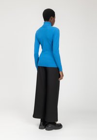 Maglione a collo alto blu con un design aderente, maniche a coste e orlo a coste, abbinato a pantaloni neri larghi e scarpe nere massicce.