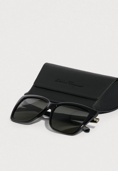 Μαύρα γυαλιά ηλίου με υπερμεγέθη γωνιακά πλαίσια και σκούρα φακούς, συνοδευόμενα από μαύρη θήκη με υφή και ανάγλυφο λογότυπο.