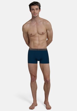 Navy blue boxer shorts with a textured fabric, featuring a dark gray elastic waistband with "Tom Tailor" logo. Model stands barefoot.
