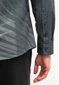 La mano di un uomo è rilassata di lato, indossando una camicia di satin verde scuro con polsini abbottonati e pantaloni neri, contro uno sfondo bianco.