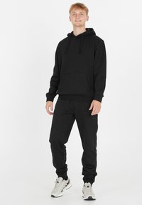 Nuori mies seisoo mustassa hupparissa ja treenihousuissa, jalassaan valkoiset ja harmaat lenkkarit valkoista taustaa vasten.