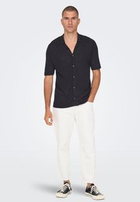 Camisa negra de manga corta hecha de tejido ligero, con botones en la parte delantera, combinada con pantalones blancos y zapatillas negras. Textura suave, corte relajado.