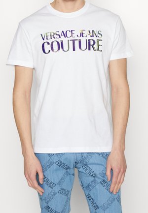 Mies, joka pukeutunut valkoiseen Versace Jeans Couture -t-paitaan ja vaaleansinisiin farkkuihin, joissa on toistuva tummansininen Versace Jeans Couture -tekstikuvio.