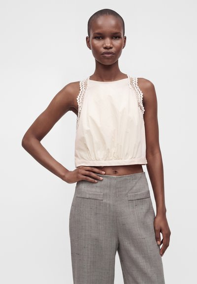 Femme portant un crop top crème sans manches avec bordure en dentelle et un pantalon gris taille haute sur mesure, debout avec une main sur la hanche sur fond blanc.
