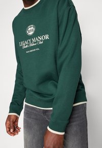 Mörkgrön sweatshirt med rund halsringning och ribbade muddar, prydd med en vit logotyp och text fram. Förstärkt med ljusare detaljer vid nederkanten.