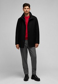 Schwarzer Mantel mit Kragen, Knopfverschluss, getragen über einem roten Pullover. Graue Hose und schwarze Schnürstiefel. Einfaches und modernes Design.