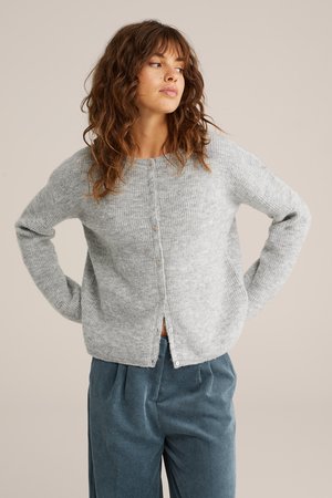 Cardigan en tricot gris clair avec un col rond, des manches longues et des boutons à l'avant. À associer avec un pantalon côtelé foncé, créant des contrastes de textures douces.