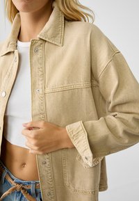 Veste en denim beige avec un col, fermeture à boutons-pression et poches latérales. Présente un tissu texturé et des manches retroussées.