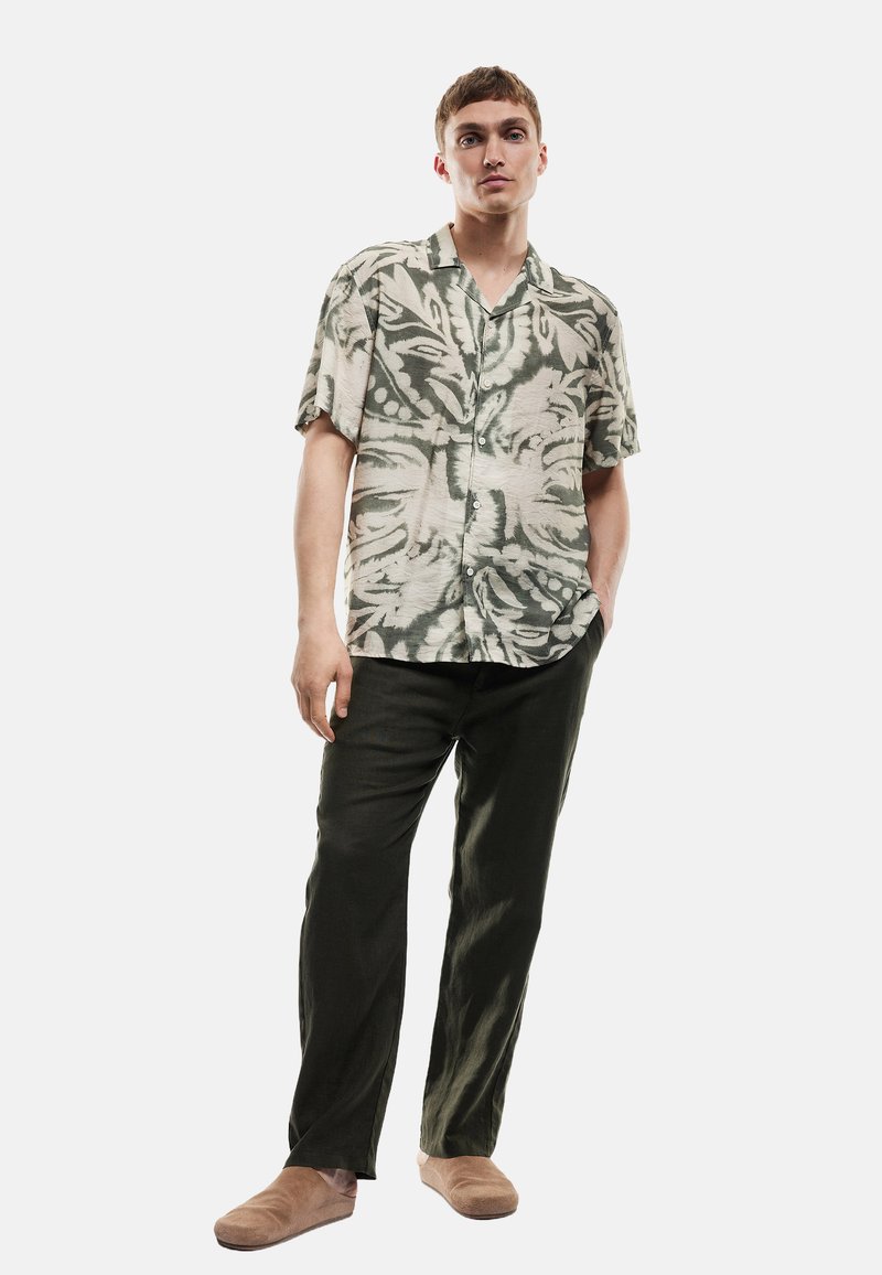 Kortebloezem, met kraag, shirt met een groen en beige bladerenpatroon, gecombineerd met donkergroene broek en bruine instap schoenen.
