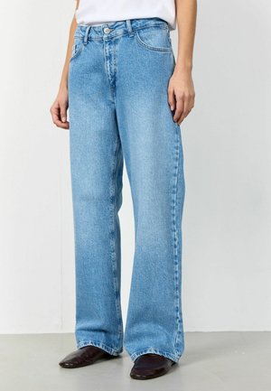 Isik kandmas helehalli laia säärega teksapükse, valget särki ja tumepruune teravatest jalanõusid, seisab heledal hallil põrandal valge seina taustal.