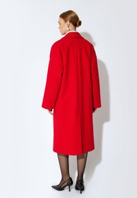 Abrigo rojo de gran tamaño con cuello con muescas, mangas largas y corte recto, combinado con zapatos de tacón alto negros y medias transparentes.