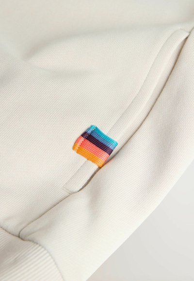 Gros plan sur un tissu crème avec une poche et une petite étiquette cousue affichant des rayures horizontales en bleu, violet, rose et orange.