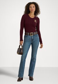 Μπορντό πουλόβερ με πλέξη καλώδιο και διακοσμητικά χρυσά κουμπιά, συνδυασμένο με μπλε τζιν παντελόνι ίσιας γραμμής και καφέ τσάντα. Το άτομο στέκεται σε ουδέτερο φόντο.