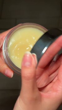 Mani che tengono e aprono un piccolo barattolo di crema o balsamo giallastro con un coperchio nero su uno sfondo scuro.