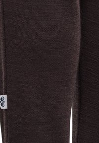 Brun stickad tyg som visar en texturerad yta, med en liten etikett som har en blå logotyp på sidan.
