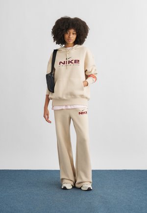 Mladá žena s kučeravými vlasmi oblečená v béžovej mikine Nike s kapucňou a nohaviciach, bielych teniskách a nesúca čiernu kabelku cez telo, stojaca na modrej podlahe.