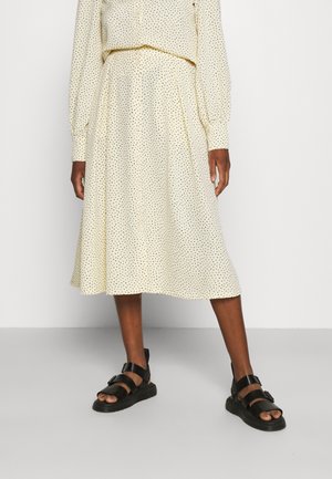 Kremowa spódnica midi w czarne groszki, noszona z dopasowaną bluzką z długim rękawem i czarnymi sandałami z klamrą na stojącej osobie.