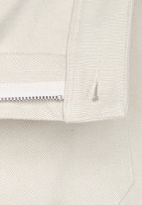 Details van een witte katoenen jas met een metalen rits, net gestikte naden en een subtiele textuur zichtbaar in de stof.