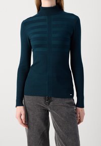 Pull à col roulé ajusté couleur sarcelle avec une texture à rayures horizontales, des poignets et un ourlet côtelés. Associé à un jean taille haute gris.