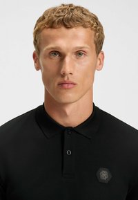 Polo noir avec un col texturé, fermeture par boutons et logo hexagonal sur la poitrine gauche. Le tissu semble lisse et ajusté.