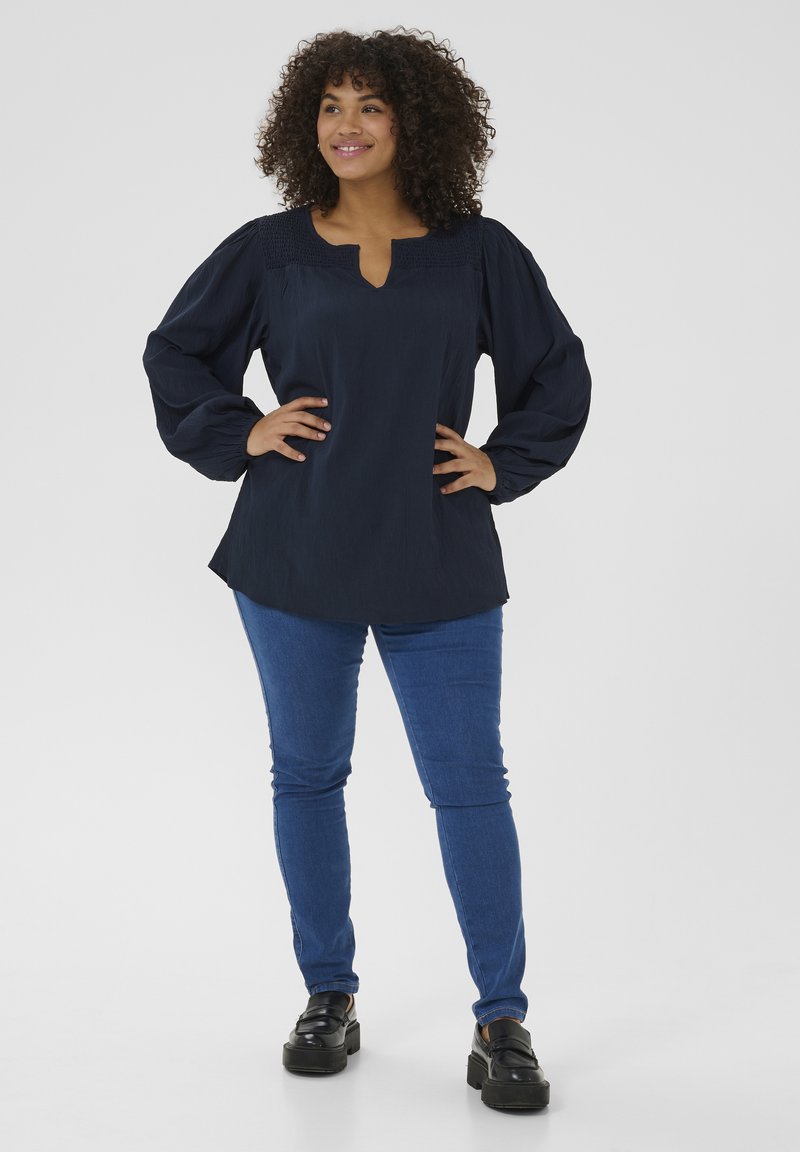 Mørkeblå bluse med smockdetaljer på skuldrene, V-hals og puffede ærmer, parret med blå tætsiddende jeans og sorte platformsko.