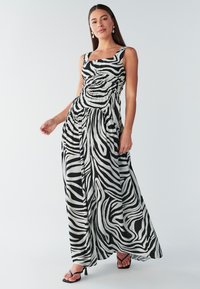 Abito lungo con stampa zebre in bianco e nero. Scollo quadrato, corpetto aderente e gonna fluente con texture a pieghe. Abbinato a sandali neri con tacco.