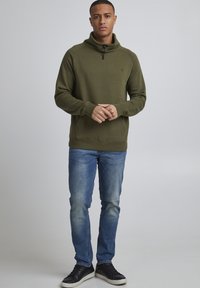 Felpa in pile verde oliva con colletto alto e mezza cerniera, con maniche lunghe e tasca a marsupio. Indossata con jeans blu e sneakers nere.