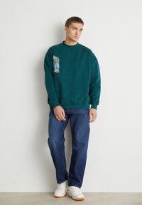 Türkise sweatshirt met een grafische patch op de linkerkant van de borst, gecombineerd met blauwe jeans en witte sneakers. De sweatshirt heeft een ronde hals.