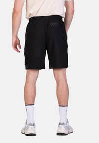 Schwarze Shorts mit einem elastischen Bund, ausgestattet mit einer Reißverschluss-Tasche auf der Rückseite und einer glatten Stofftextur. Getragen mit weißen Socken und Turnschuhen.