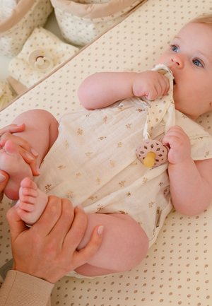 Μωρό που ξαπλώνει σε πολύχρωμο στρώμα αλλαγής, κρατώντας και μασώντας ένα ύφασμα προσκολλημένο σε πιπίλα, ενώ ενήλικα χέρια κρατούν απαλά τα πόδια του μωρού.