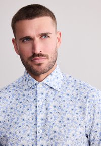 Mladý muž s krátkymi vlasmi a bradou, oblečený v zapnutej svetlomodrej košeli s bielym kvetinovým vzorom, mierne sa pozerá doprava.